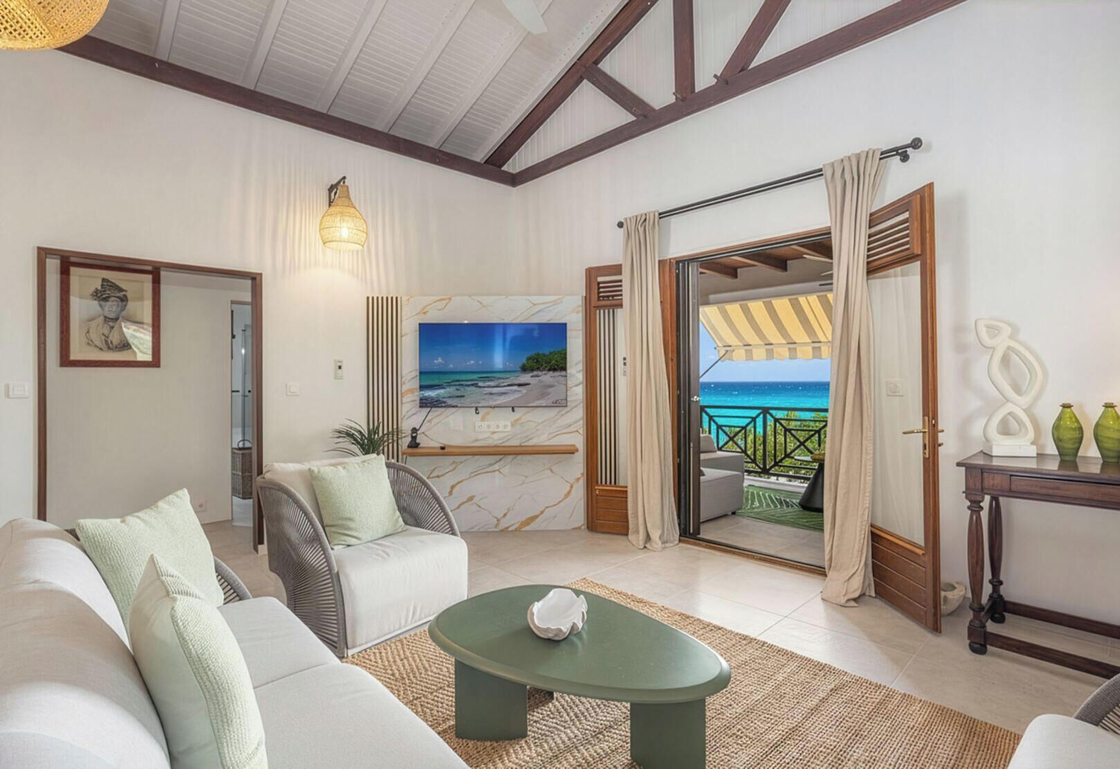 A living room with a sofa, armchair, and coffee table, a TV mounted on the wall, and a view of the beach through an open door.