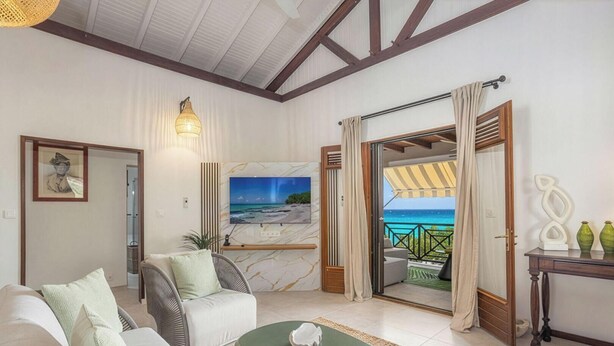 Een woonkamer met een bank, fauteuil en salontafel, een aan de muur bevestigde televisie en uitzicht op het strand door een geopende deur.