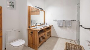 Een badkamer met een houten wastafelmeubel, twee wastafels, een grote spiegel, een toilet, een douche en een rieten mand.