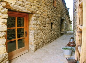 A stone building with a wooden door and window, a narrow pathway, and metal containers on the ground. - Isolated stone and timber house in the forest (Lleida)
