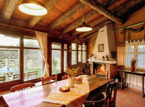 A rustic interior with a wooden dining table, chairs, a fireplace, and large windows. - Isolated stone and timber house in the forest (Lleida)