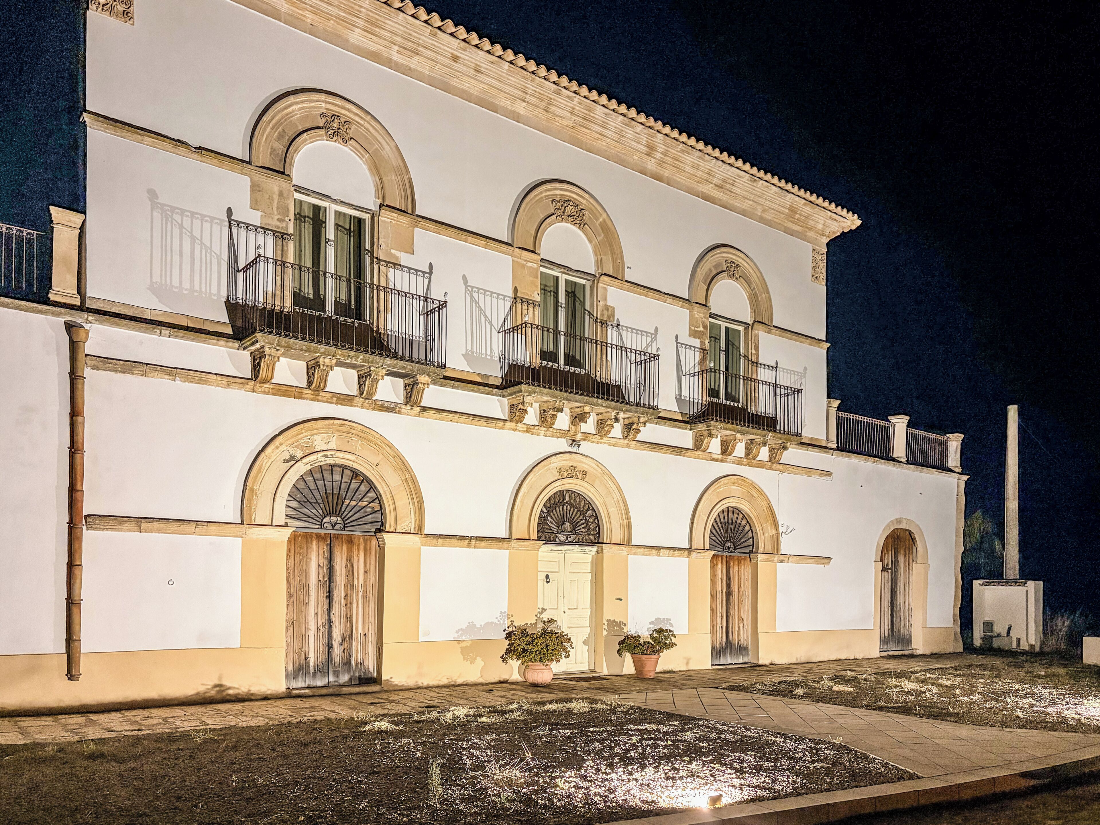 A two-story building with arched windows and balconies, a gated entrance, and potted plants.