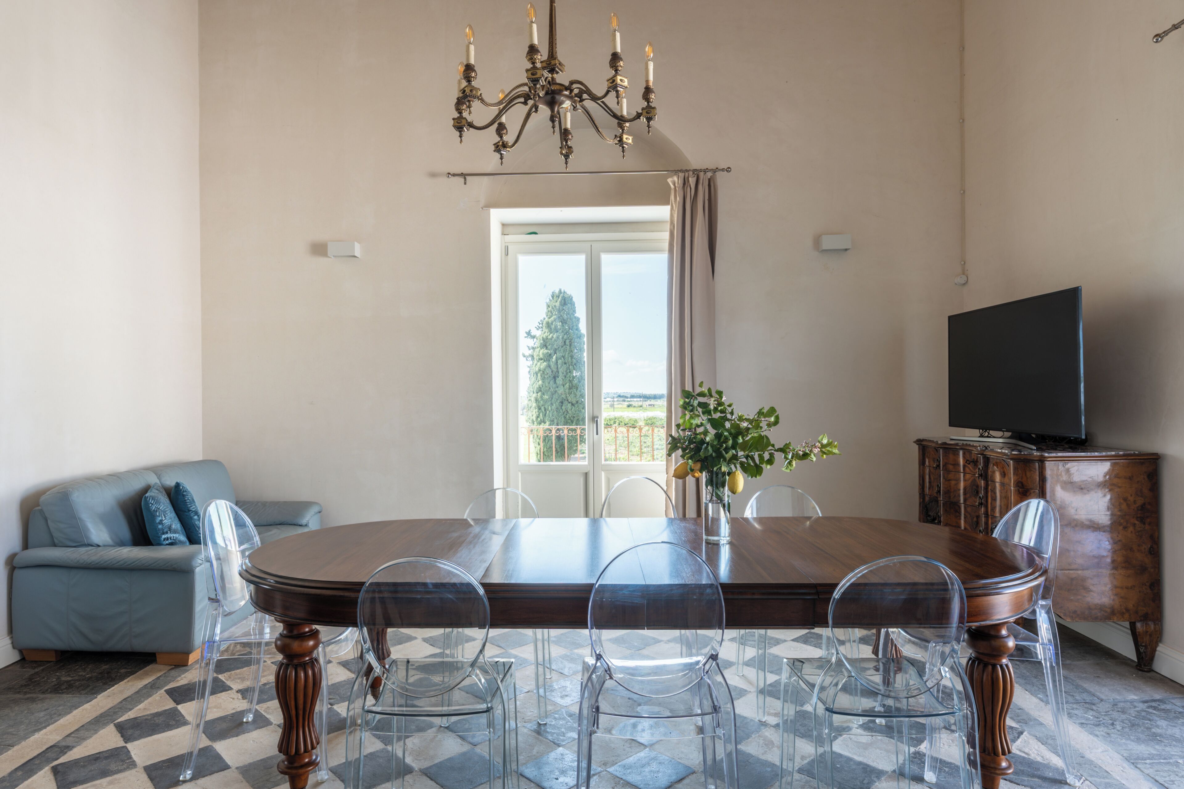A dining area with a wooden table, clear chairs, a chandelier, a television, and a sofa.