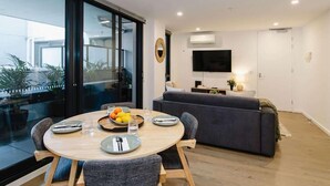 A modern living room with a dining table, grey sofa, and a flat-screen TV mounted on the wall. - 101-251 Johnston St (Abbotsford)