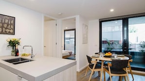 A modern kitchen with a central island, a dining area with wooden chairs, and a large sliding glass door leading to an outdoor area. - 101-251 Johnston St (Abbotsford)
