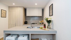 A modern kitchen with a central island, bar stools, a wine bottle, and a vase with flowers. - 101-251 Johnston St (Abbotsford)