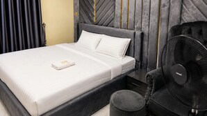 A neatly made bed with white linens, a black upholstered headboard, a black chair with a fan, and a wall-mounted air conditioner. - Rama Garden Hotel (Akure)
