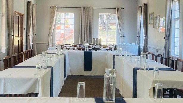 A dining area with tables set for service, large windows with curtains, and a view of the outdoors.