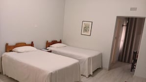A room with two single beds, a wooden bench, a framed picture on the wall, and a door leading to a corridor.