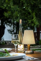 Uma garrafa de vinho, um copo e um abajur sobre uma mesa externa, com vegetação ao fundo.