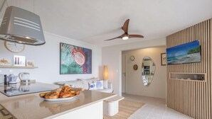 A modern kitchen with a central island, a ceiling fan, a large painting, and a TV mounted on a wooden panel wall. - Éole, a charming villa, within walking distance of the beach, with a view of the sea (Sainte-Luce)