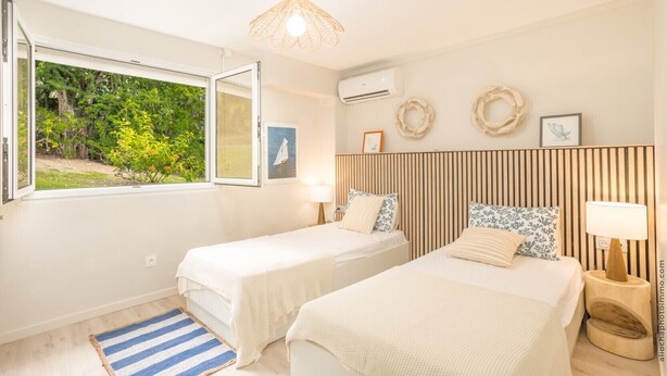 A bedroom with two beds, a window with a view of greenery, an air conditioning unit, and a wall-mounted lamp. - Éole, a charming villa, within walking distance of the beach, with a view of the sea (Sainte-Luce)
