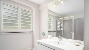 A bathroom with a large mirror, white vanity, and a window with blinds. - Éole, a charming villa, within walking distance of the beach, with a view of the sea (Sainte-Luce)