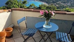 Un balcone con tavolo e sedie, affacciato su un paesaggio costiero con vista sul mare e colline in lontananza.