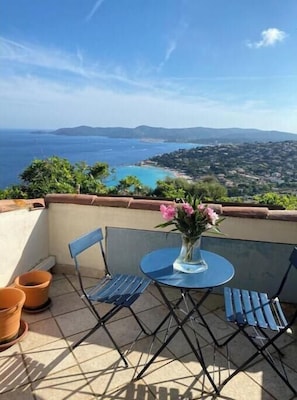 Ein Balkon mit Tisch und Stühlen, der auf eine Küstenlandschaft mit Blick auf das Meer und ferne Hügel blickt.