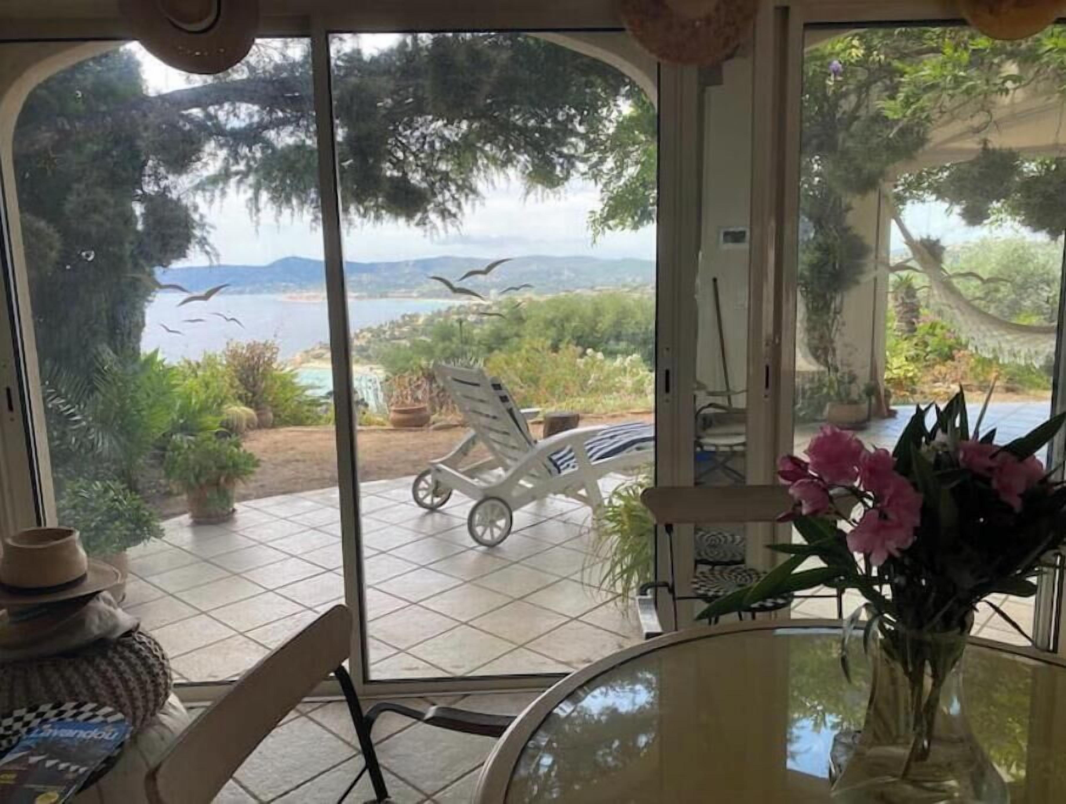 Una habitació amb vista a un jardí i a l'aigua, una taula amb un ram i una cadira.