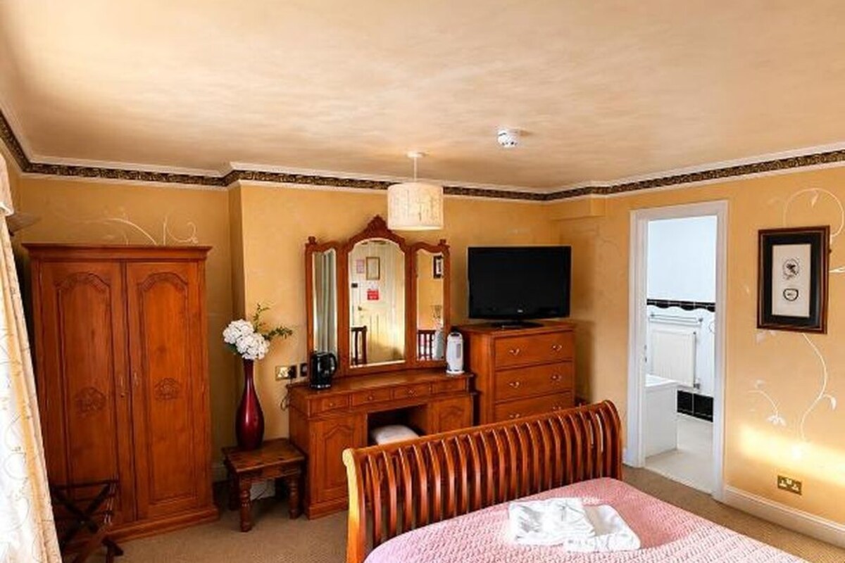 A bedroom with a bed, a dresser, a mirror, a television, and a door leading to a bathroom.