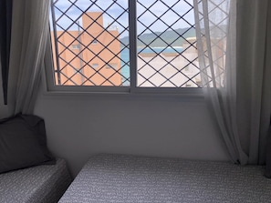 Ein Zimmer mit Bett, einem Fenster mit Blick auf Gebäude und einen Strand, sowie Vorhängen.