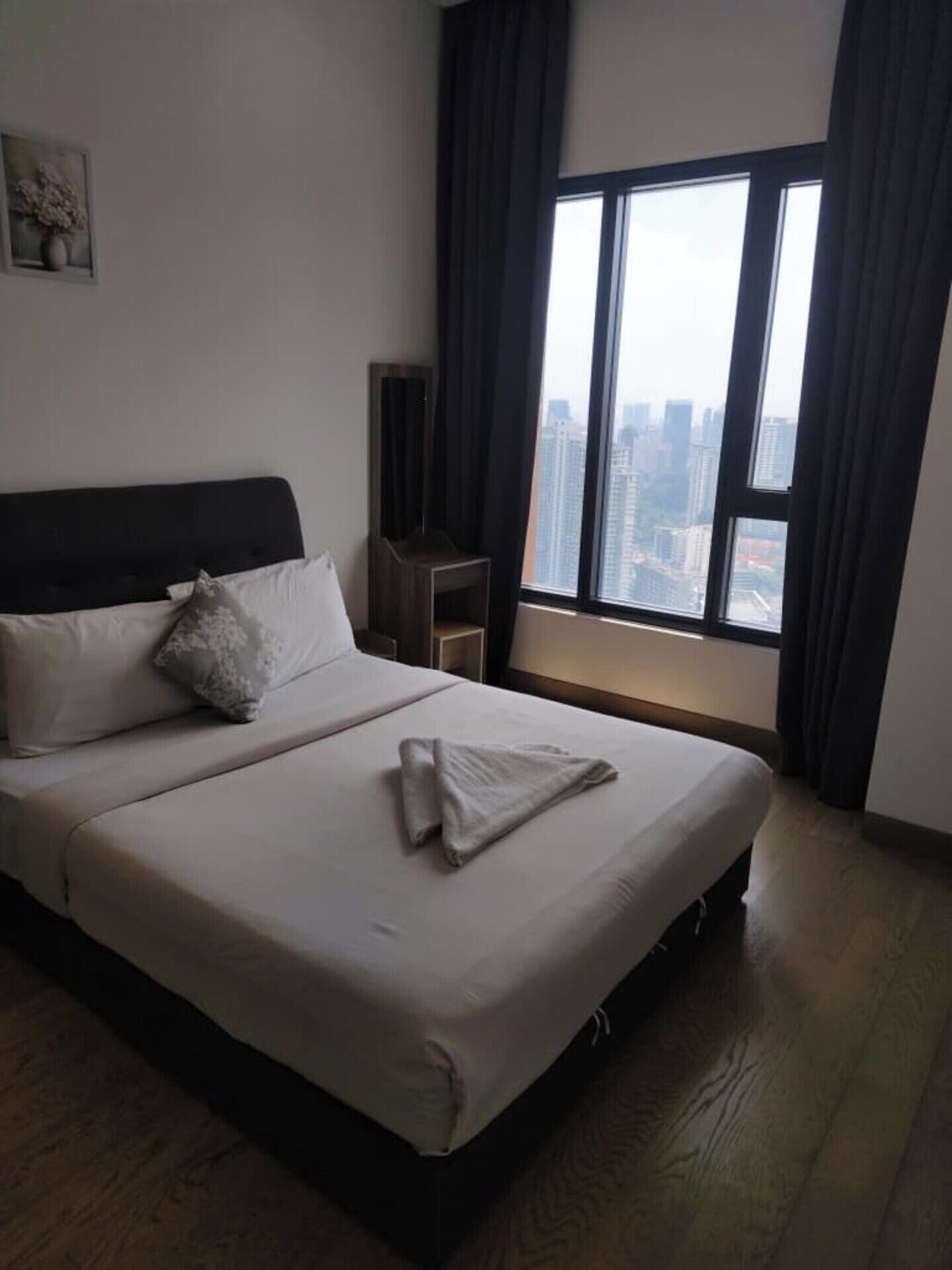 A hotel room with a large bed, a view of the city through a window, and a small table with a chair.