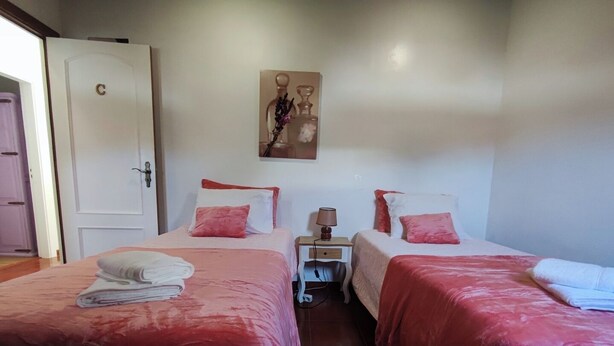 Une chambre d’hôtel avec deux lits, chacun recouvert d’une couverture rouge et doté d’oreillers blancs, une table de chevet avec une lampe, et une porte marquée d’un « C ».