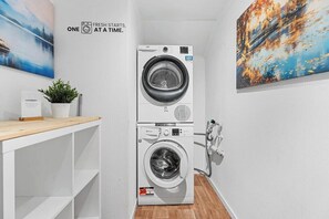 A laundry room with two stacked front-loading washing machines, a wooden shelf, a potted plant, and a painting on the wall.
