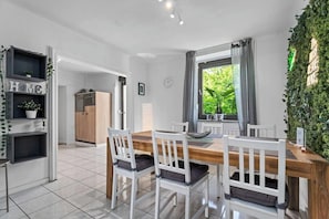 A modern kitchen with a wooden table, white chairs, and a view of greenery outside.