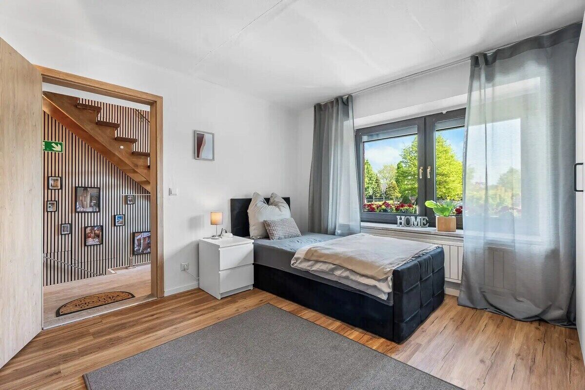 A bedroom with a bed, bedside table, window with curtains, and a view of greenery outside.