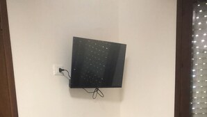 Una habitación pequeña con un escritorio blanco, una silla negra, un refrigerador blanco y un televisor montado en la pared.