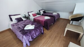 Een kamer met drie bedden, elk voorzien van een paarse sprei, witte kussens en een witte handdoek. Er staat een klein houten tafeltje en een stoel.