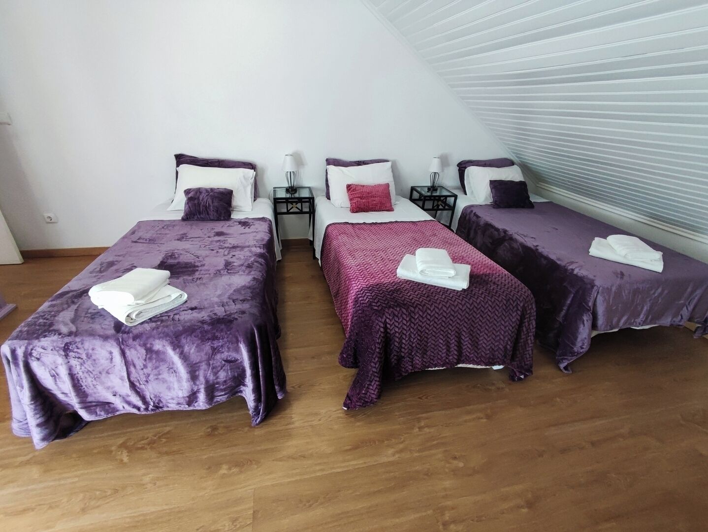 Une chambre avec trois lits, chacun recouvert d’une couverture violette et agrémenté d’oreillers blancs, et une petite table placée entre eux.