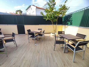 An outdoor patio area with multiple tables and chairs, a small tree, and a green fence. - Lisbon Airport GuestHouse Gal - Suite w/ Wc (Lisboa)