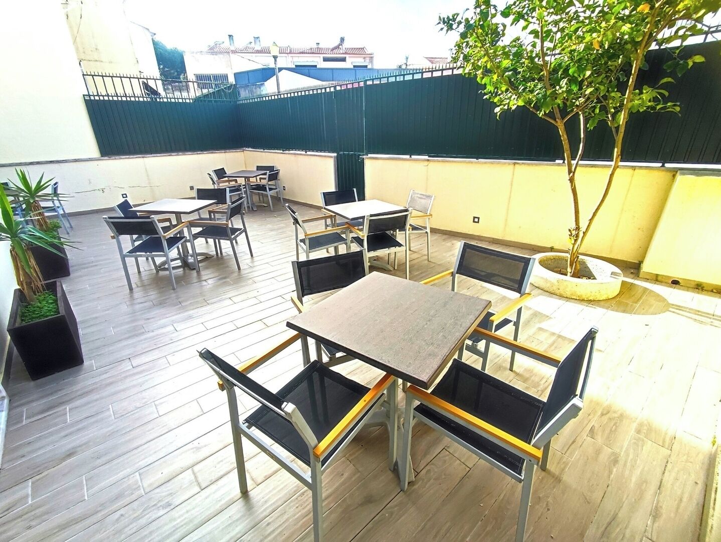 Un espacio al aire libre con mesas y sillas, un árbol pequeño y vistas de edificios al fondo.