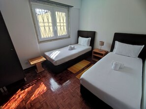 A hotel room with a single bed, a smaller bed, a window with metal bars, a bedside table with a lamp, and a bench. - Lisbon Airport GuestHouse Gal – Twin Bedroom (Lisbon)
