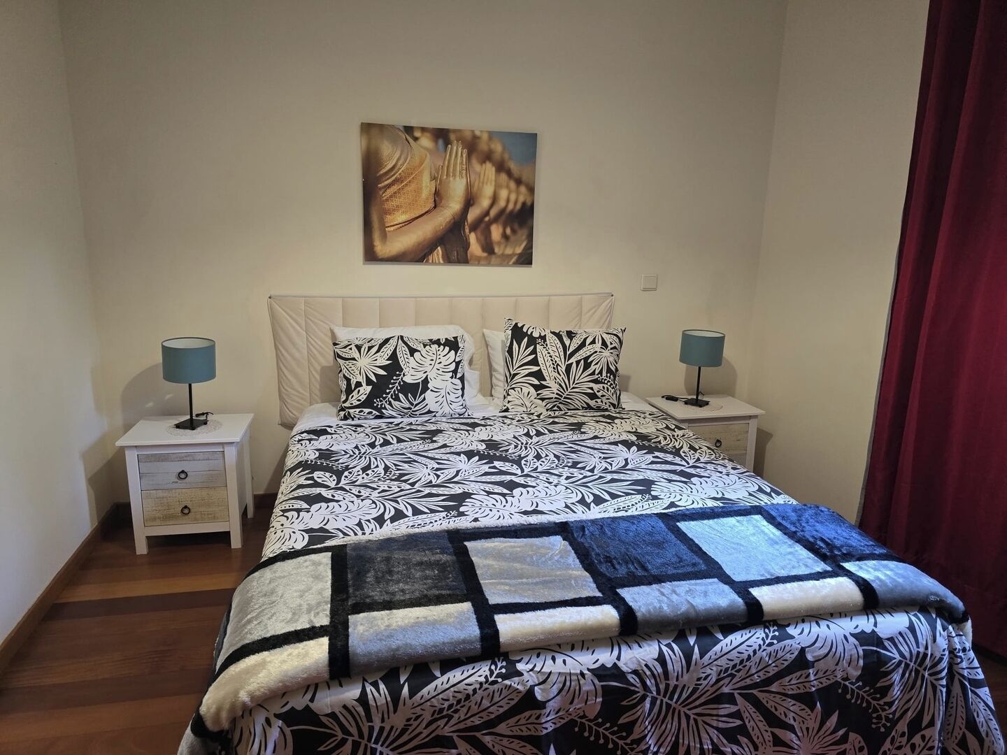 Un dormitorio con cama, dos mesitas de noche, una lámpara de noche, una manta y un cuadro en la pared.