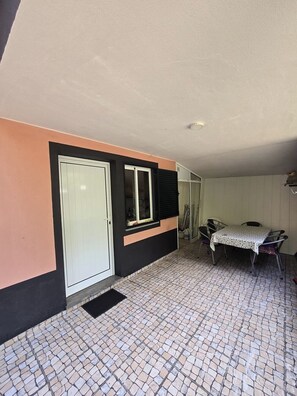 A room with a tiled floor, a white door, a window with blinds, a table with chairs, and a small shelf. - Madeira, 2 Bedroom Nature Retreat near Santa Cruz (Gaula)