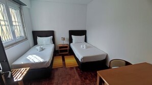 Quarto pequeno e limpo com duas camas de solteiro, uma mesa de madeira e uma janela com grades.