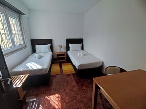 A small, clean room with two single beds, a wooden table, and a window with bars. - Lisbon Airport GuestHouse Gal – Twin Bedroom (Lisbon)