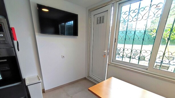 Quarto pequeno com televisão fixada na parede, uma porta, uma janela com grades de metal, uma mesa de madeira e um banco.