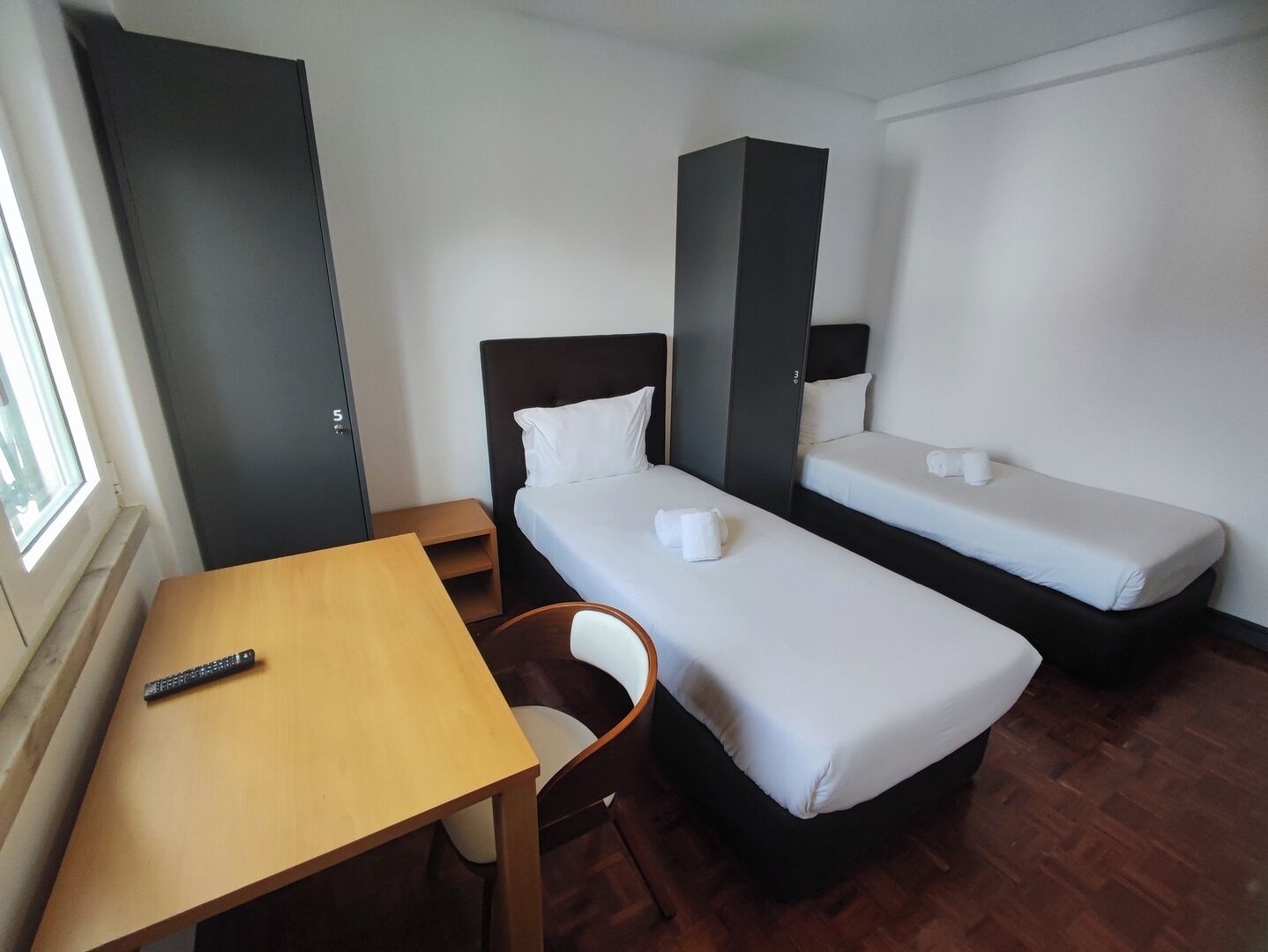 Habitación de hotel con dos camas individuales, un escritorio de madera, una silla y una mesita.