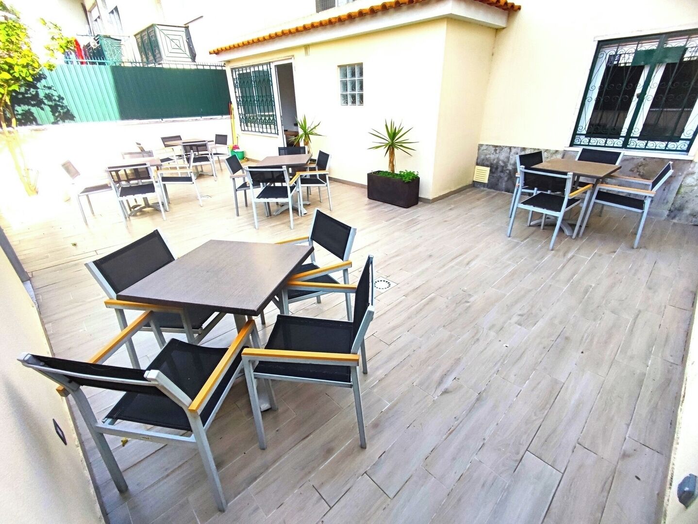 Un patio exterior con mesas y sillas, una planta en maceta y un edificio con techo de tejas.