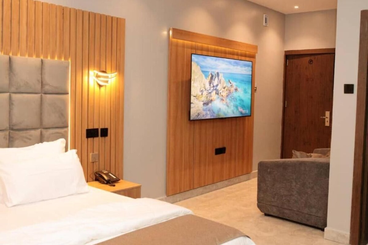 Een moderne hotelkamer met een groot bed, een televisie bevestigd aan een houten wandpaneel, een grijze bank en een houten deur.