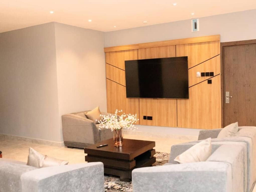 A modern living room with a flat-screen TV mounted on a wooden wall, a sofa, a coffee table with a vase, and a door to the right.