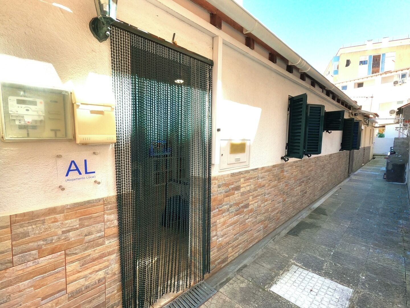 A narrow alleyway with a building featuring a metal gate, a sign reading 'AL (Alojamento Local)', and a brick wall.