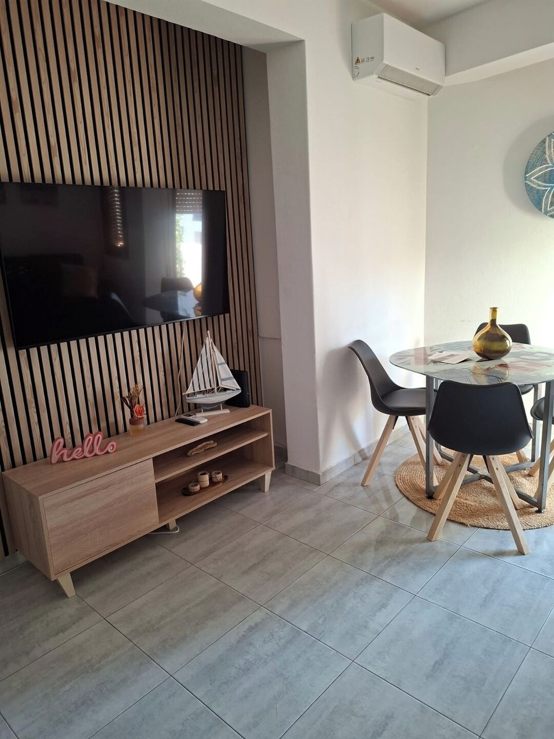 A modern living room with a flat-screen TV mounted on a wooden panel wall, a wooden entertainment unit with a 'hello' sign, a round dining table with chairs, and a woven rug.