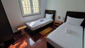 Habitación de hotel con una cama individual, una cama más pequeña, una ventana con barrotes, una mesita de noche con lámpara y un banco.