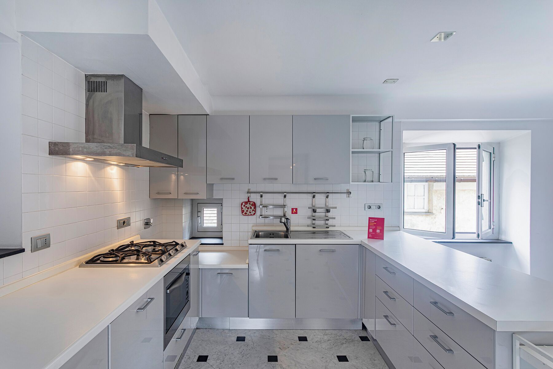 Une cuisine moderne avec des meubles blancs, une cuisinière à gaz, un four encastré et une fenêtre avec des stores.