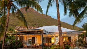 A modern house with a covered patio, surrounded by lush greenery and palm trees, with a mountain in the background. - Luxury Beachfront Villa • Private Pool, Beach, AC, Calm Lagoon (La Preneuse)