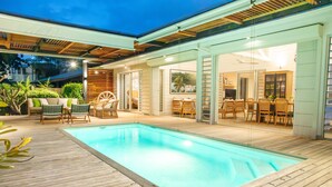 A swimming pool with a wooden deck, surrounded by wicker furniture and a glass door leading to an indoor area with a dining table and chairs. - Luxury Beachfront Villa • Private Pool, Beach, AC, Calm Lagoon (La Preneuse)