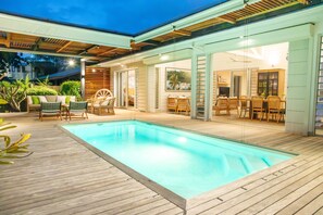 Ein Schwimmbecken mit Holzterrasse, umgeben von Korbmöbeln und einer Glastür zu einem Innenbereich mit Esstisch und Stühlen.