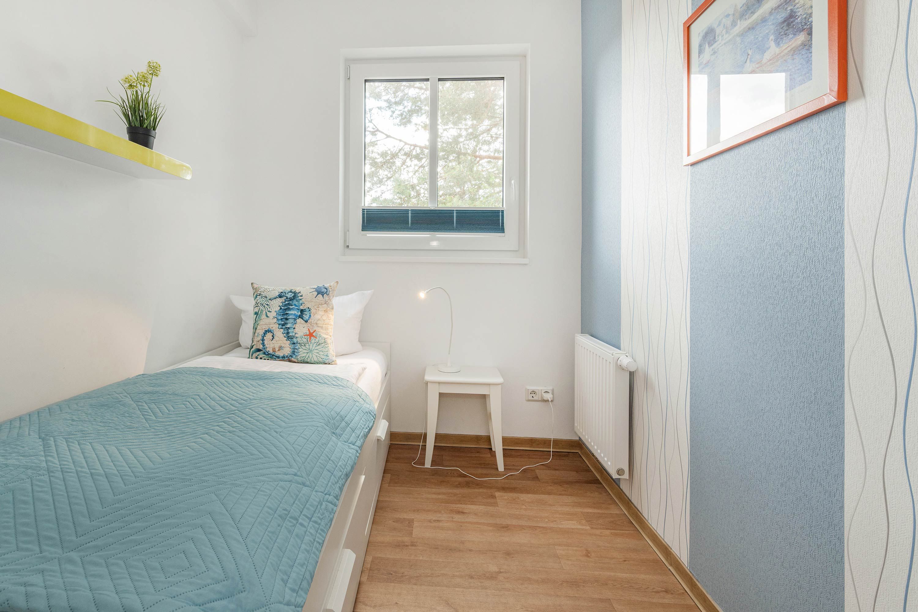 Ein Schlafzimmer mit Bett, Nachttisch, Fenster, Heizkörper und einer Wand mit einem Bilderrahmen.